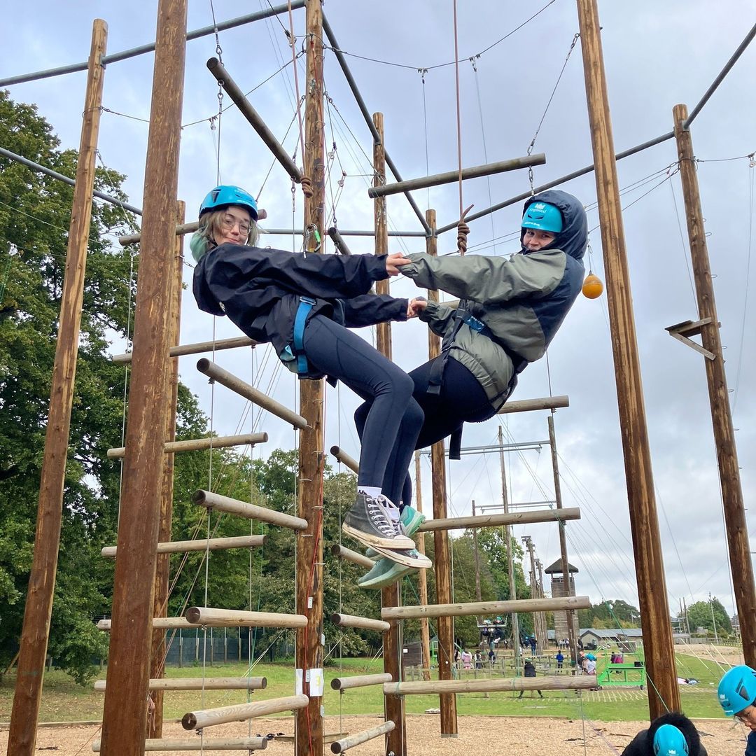 A photo of two young students on an NCS experience wearing safety helmets and harnesses at a high ropes adventure course. They are suspended in the air, holding each others hands whilst looking sideways to the camera. The structure consists of vertical wooden poles and connecting ropes. In the background, there is a view of the park with more of the course and other participants preparing for the activity.
