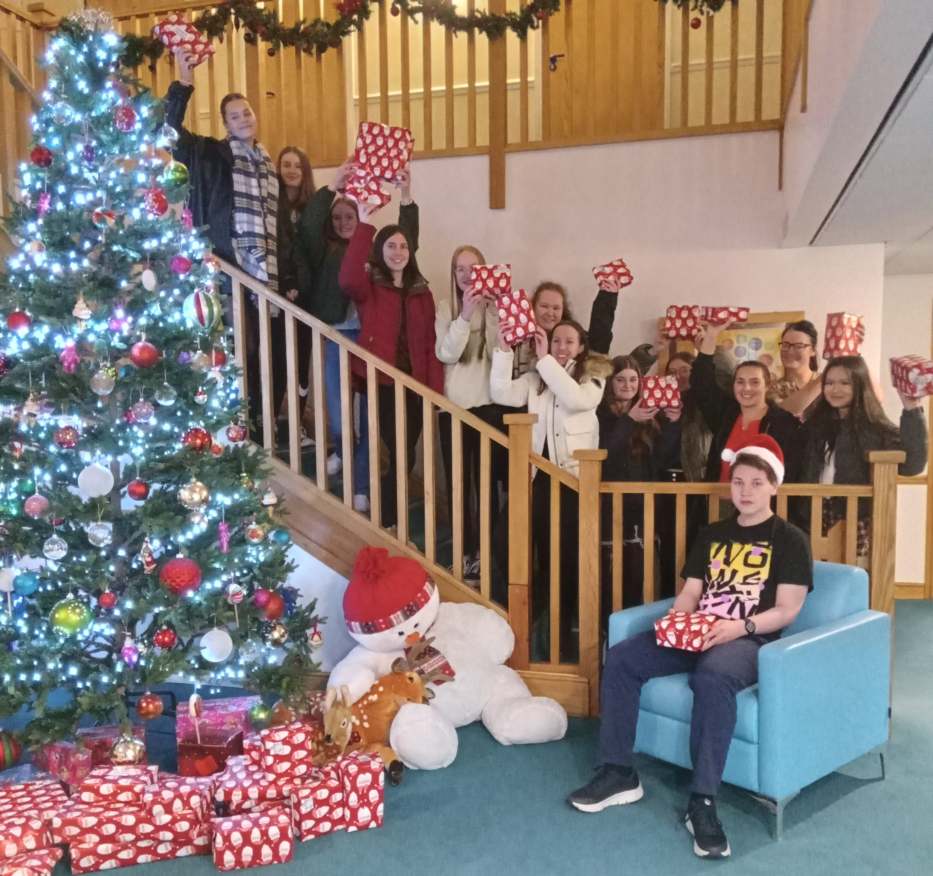 A group of young people standing on the staircase happy with a christmas tree and gifts wrapped around the tree
