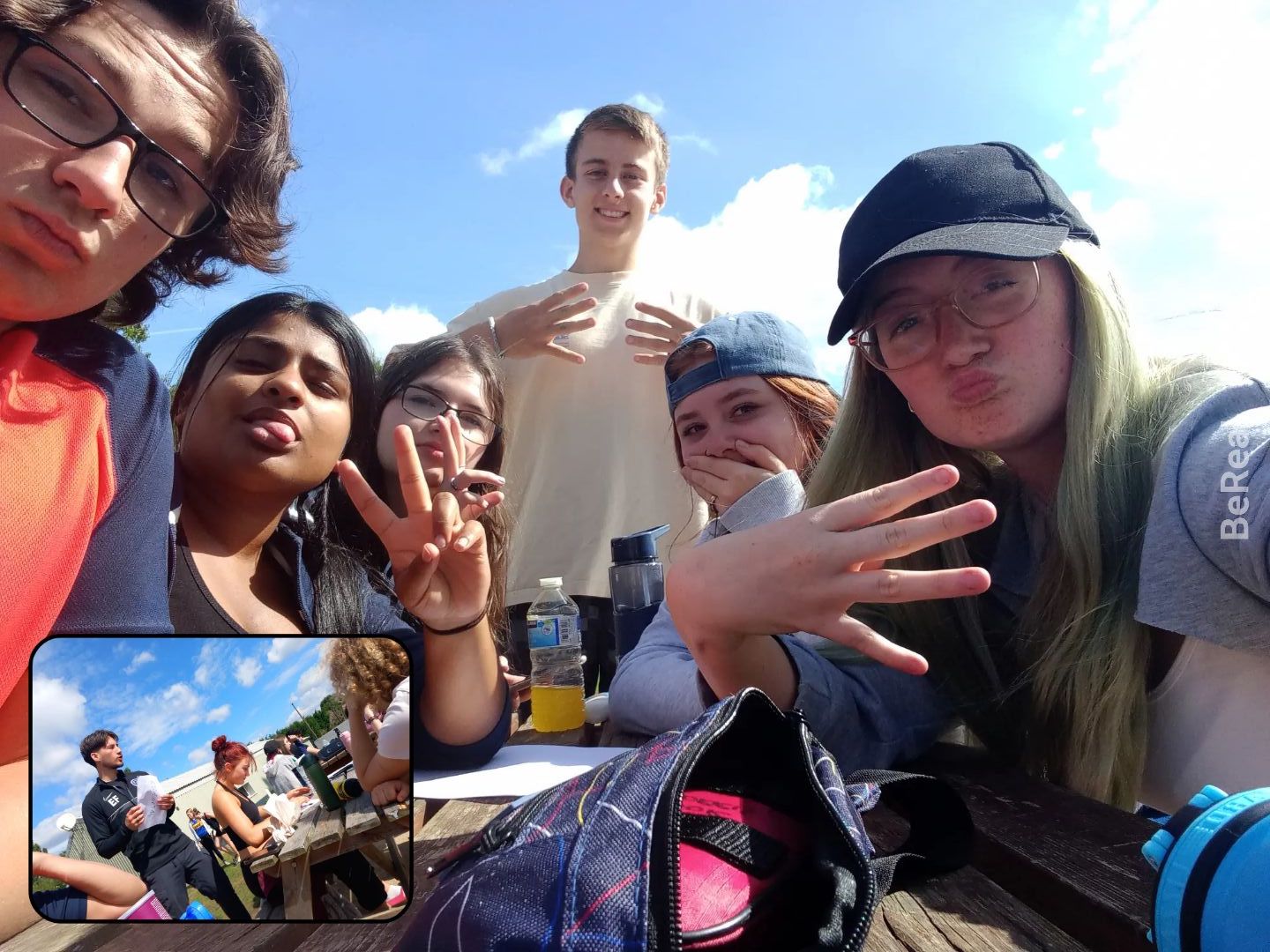 Charlotte is taking a selfie on the BeReal app. She's at the front wearing a cap and holding four fingers up to the camera, and her friends next to her are also doing peace signs with their hands. 