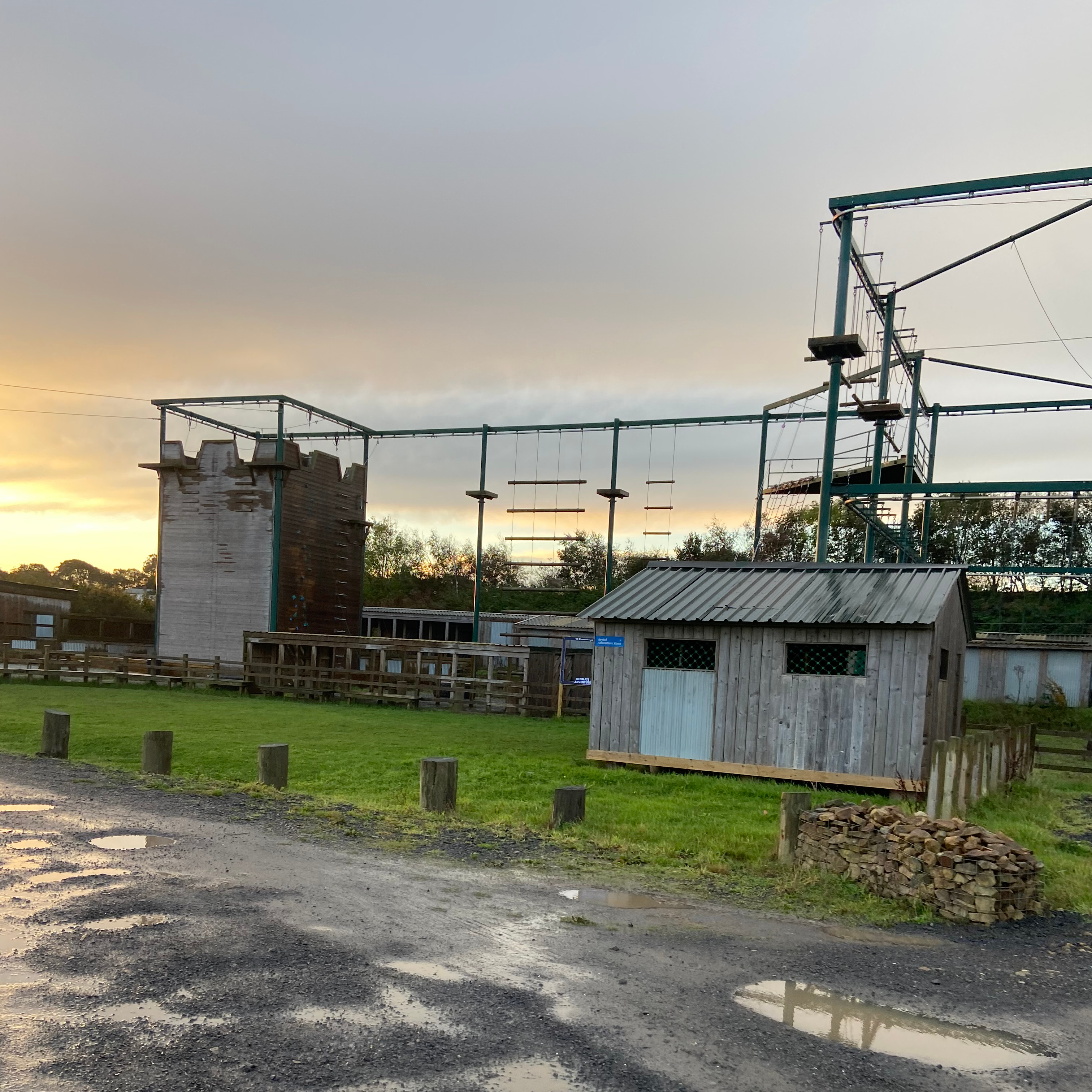 An empty high ropes course with a wooden hut, grass and some puddles in front of it. In the background the sun is setting. 
