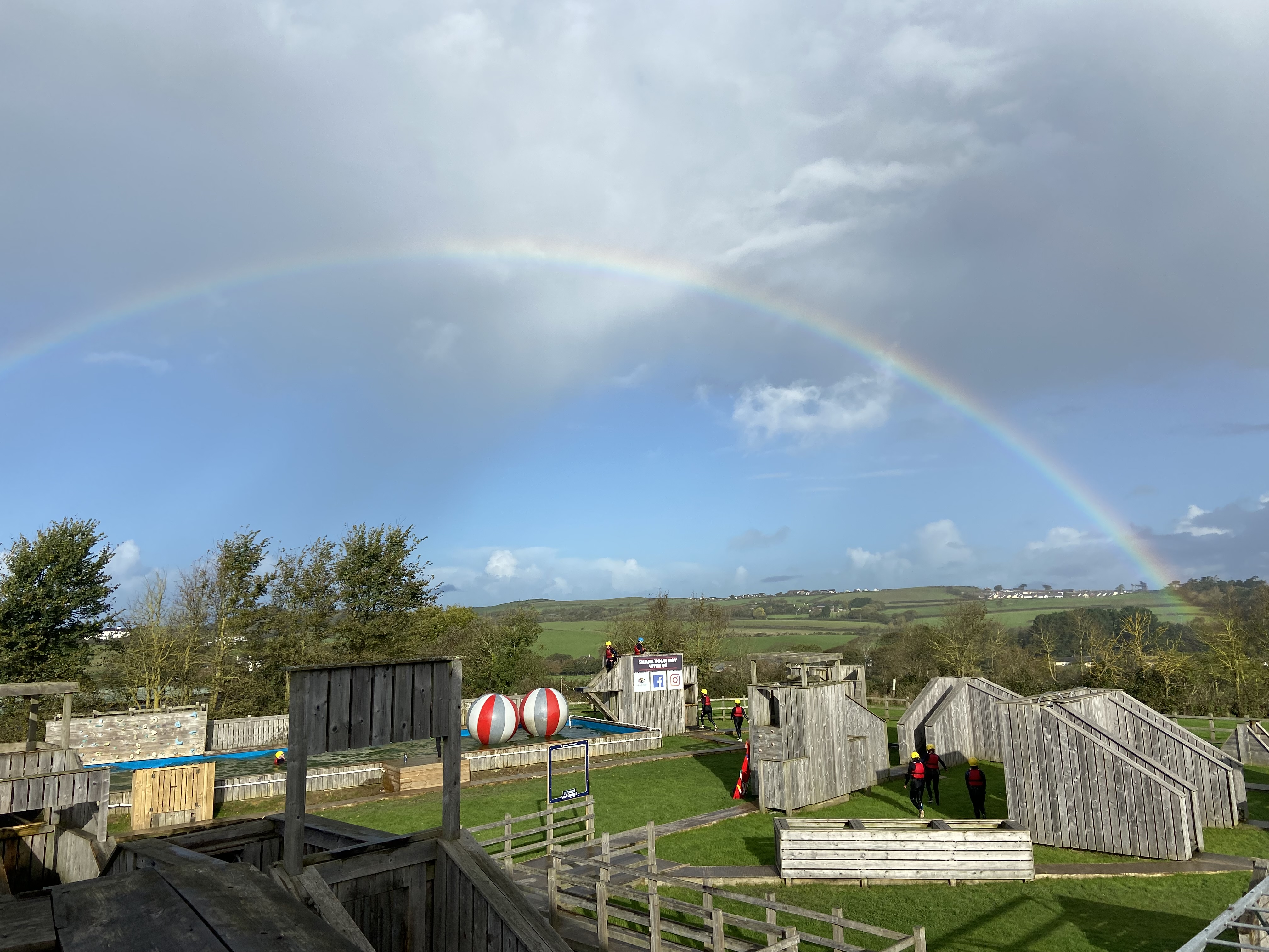 A rainbow in the sky sits over an ultimate assault course. The course features wooden wall climbs and a water challenge with giant inflatable balls.