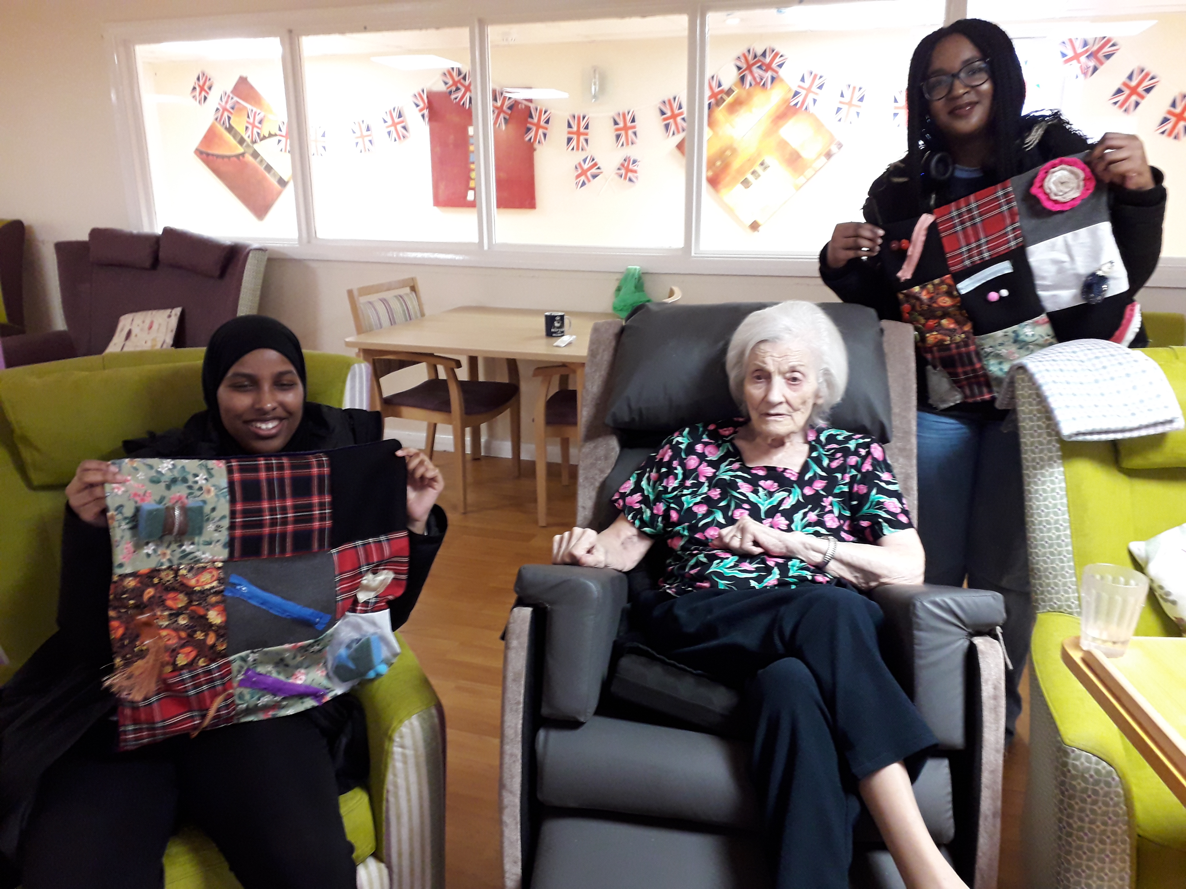 Two ladies holding a hand made blanket with an elderly woman on a chair for the Dementia Fidget Blanket Project