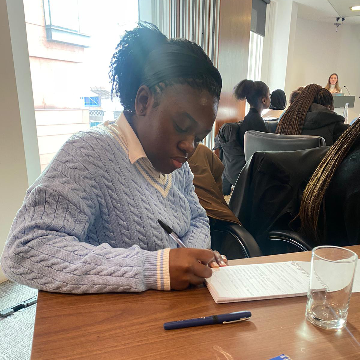 A picture of Jemimah, a young woman seated in a conference room with other participants in the background, writing notes in a notebook. She is wearing a blue cable knit sweater over a white collared shirt and has braided hair. A glass of water and a blue pen are on the table beside her, and in the background, a white woman is giving a presentation.