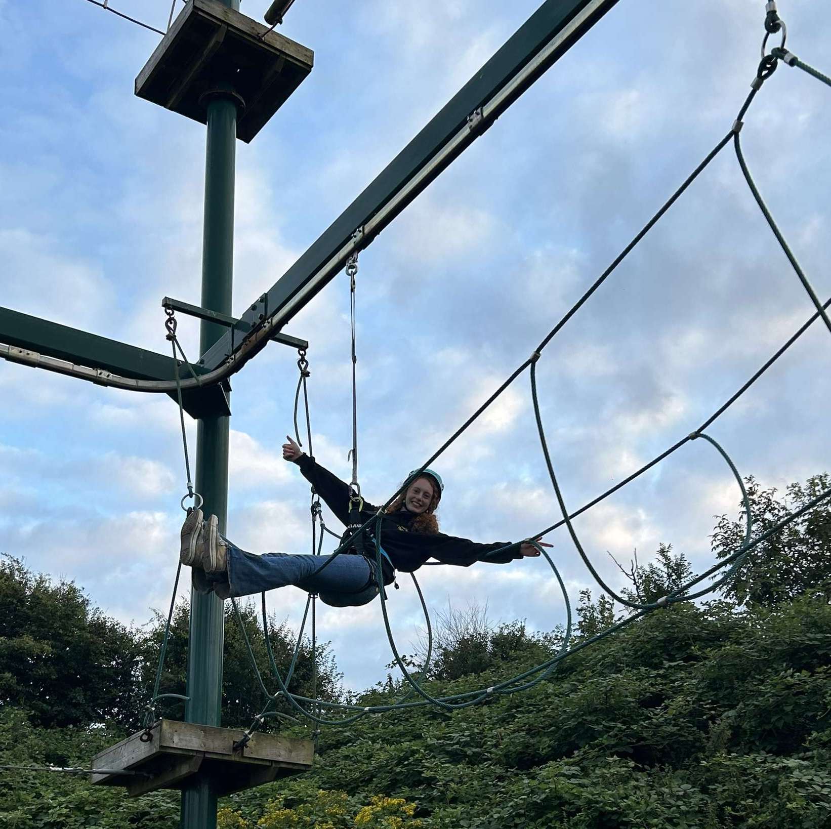  Lilla, a white young person, sits mid-air with her legs in front of her and her arms out wide and a big smile on her face on a high ropes course. She is attached with a safety rope which is fastened across her middle as well as also wearing a helmet