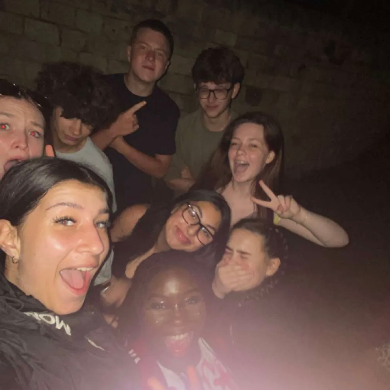 A group of young individuals standing close, smiling, and posing for a photograph, with some doing the peace sign capturing a moment of togetherness.