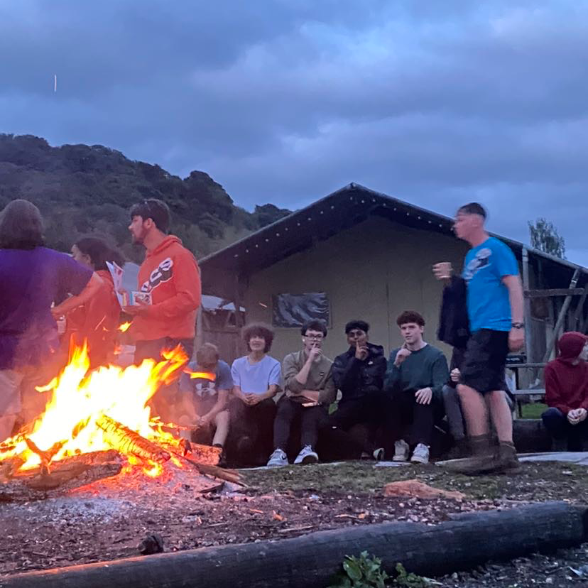A group of young boys seated around a campfire, exchanging tales and creating lasting moments while some of them wear NCS shirts enjoying the NCS experience.