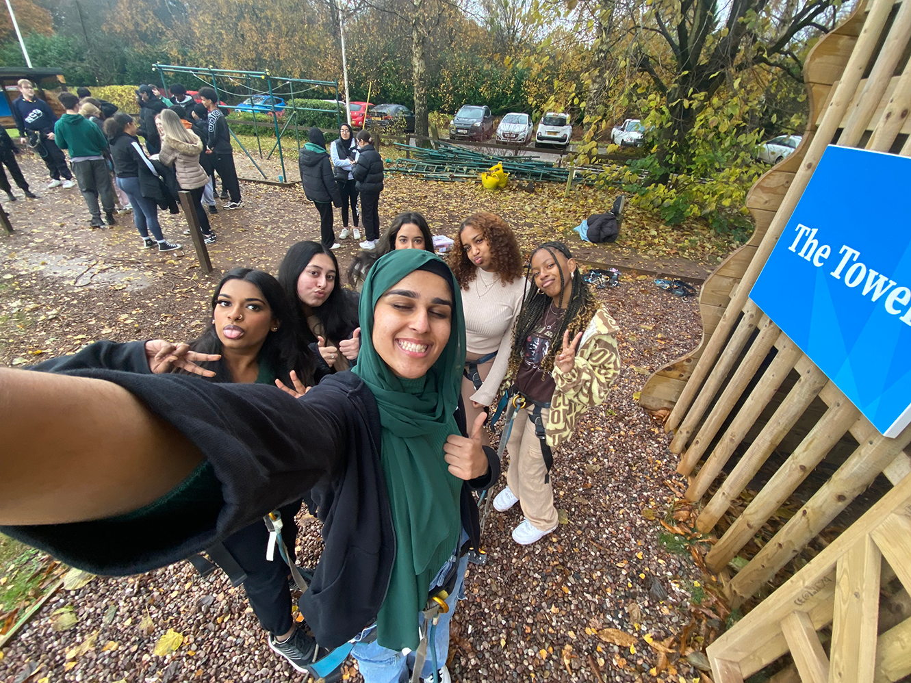 A young woman wearing a head scarf is taking a selfie with Rihanna and four other friends outside. They’re standing close to a tower assault course.