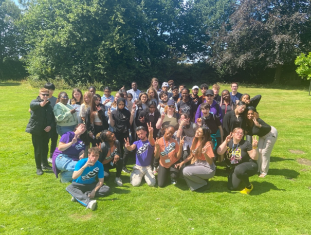Rihanna and her NCS away from home group grouped together for a photo on green grass. Some people are wearing NCS t-shirts and doing a 'peace sign' with their hands.