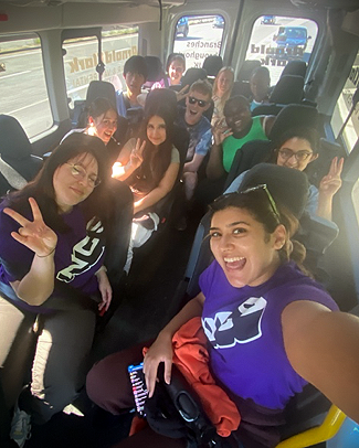 A Team Leader with an NCS t-shirt on is taking a selfie from the front of the bus. Rihanna and her friends are seated behind, and doing peace hand signs. 