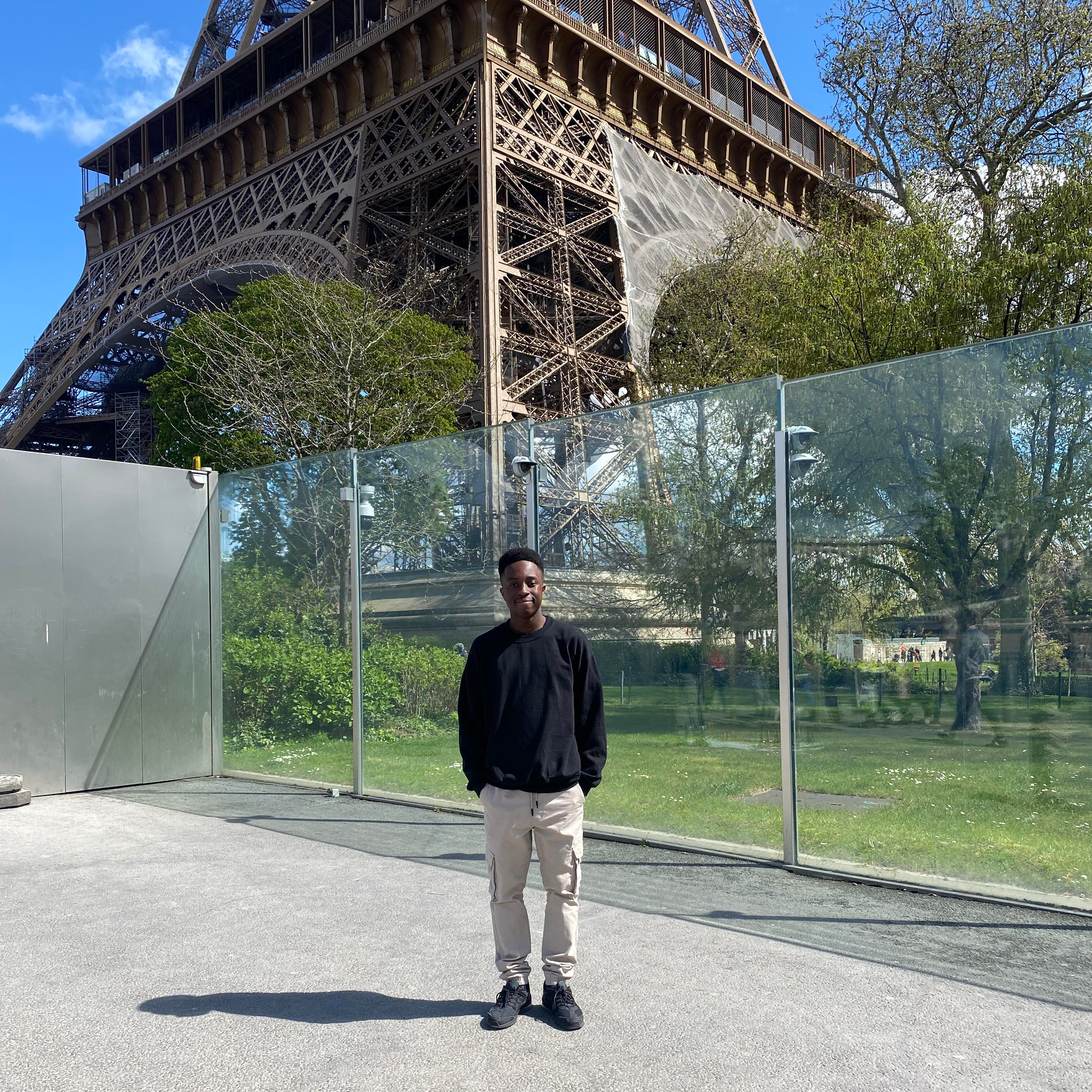 Ryan a young black man wearing a black shirt and grey jeans is standing in front of the Eiffel Tower in Paris