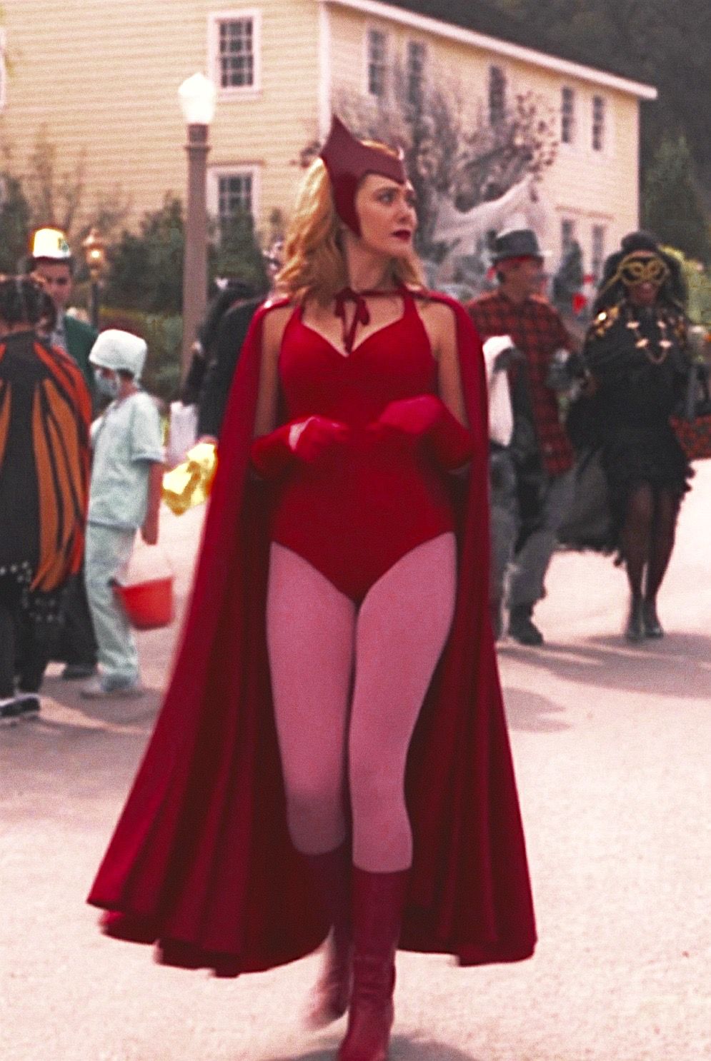 Elizabeth Olsen as Wanda Maximoff, she is walking down the street wearing a red leotard, cape and gloves, with pink tights and a red crown. Behind her are other people dressed up for Halloween.