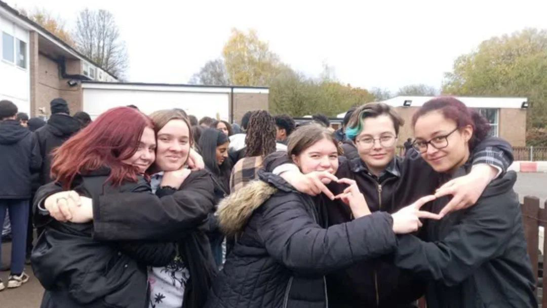 A group of young women are hugging in the school playground. The group of three on the right are forming heart shapes with their fingers. 