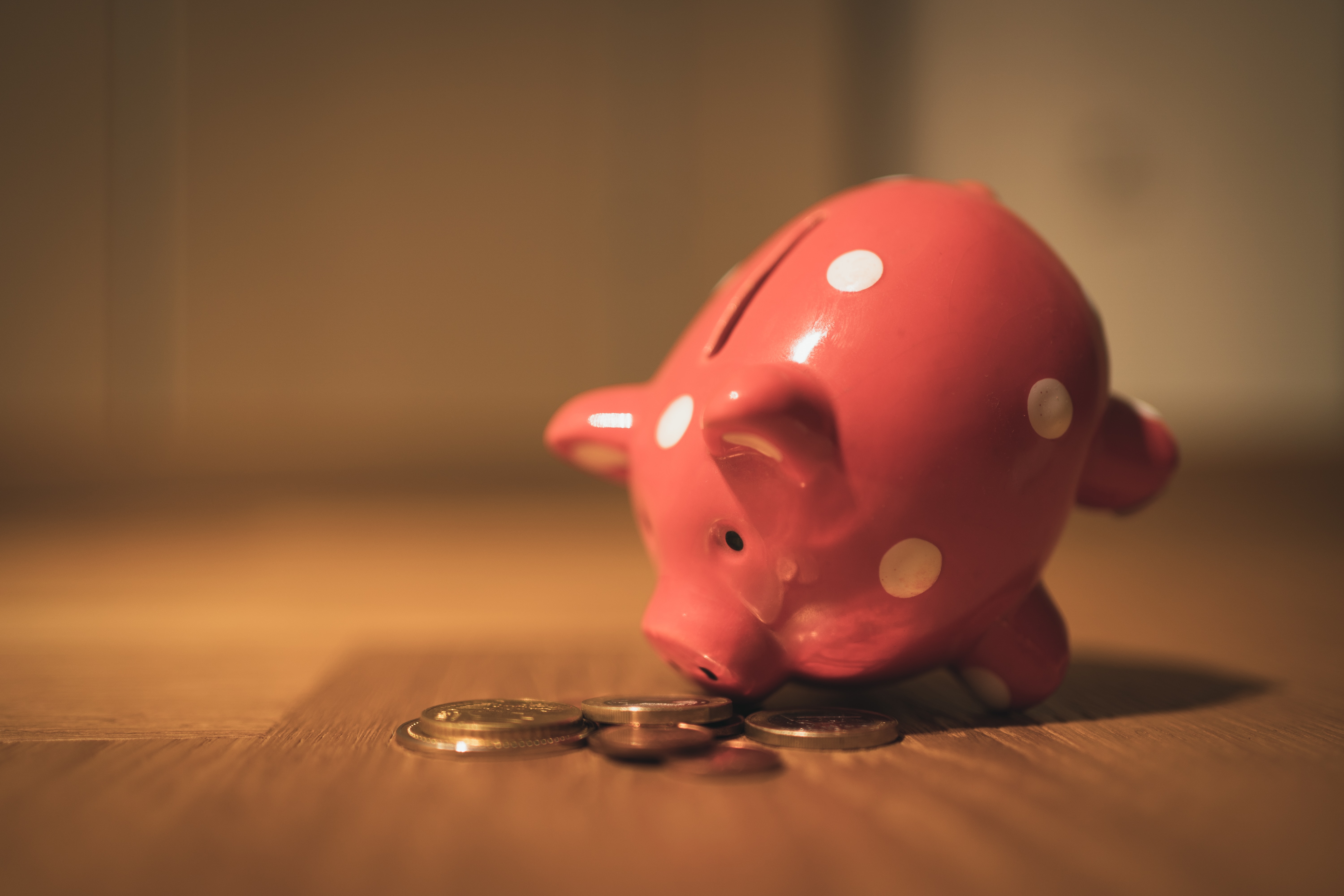 A pink piggy bank with white dots on it sat on a wooden table, the piggy bank is perched on its front legs with the snout touching a pile of coins. 