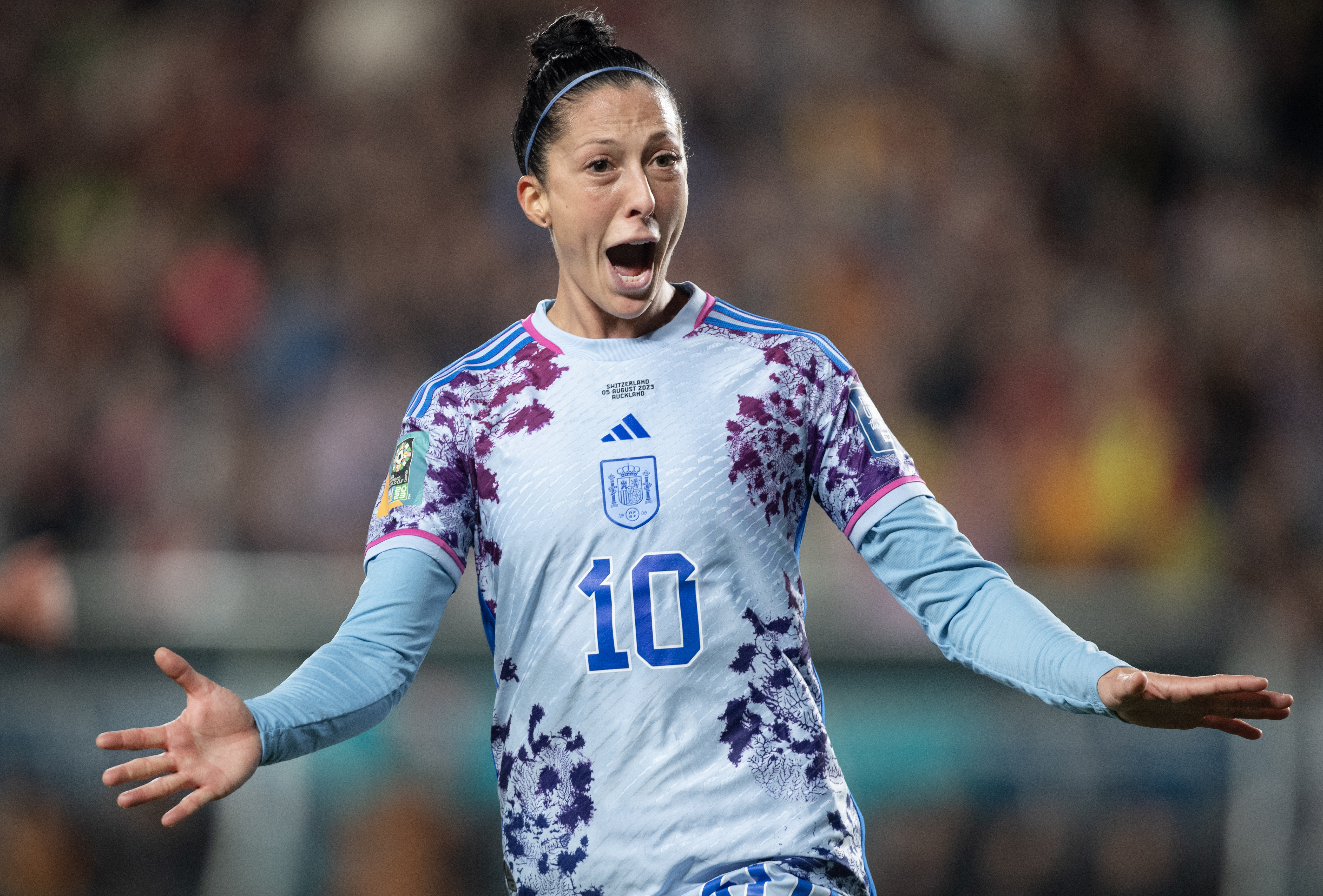 Jenni Hermoso a female soccer player in action, wearing a blue and purple patterned jersey with the number 10. She has her mouth open as if shouting and her arms extended to the side, expressing intense emotion during a game. 
