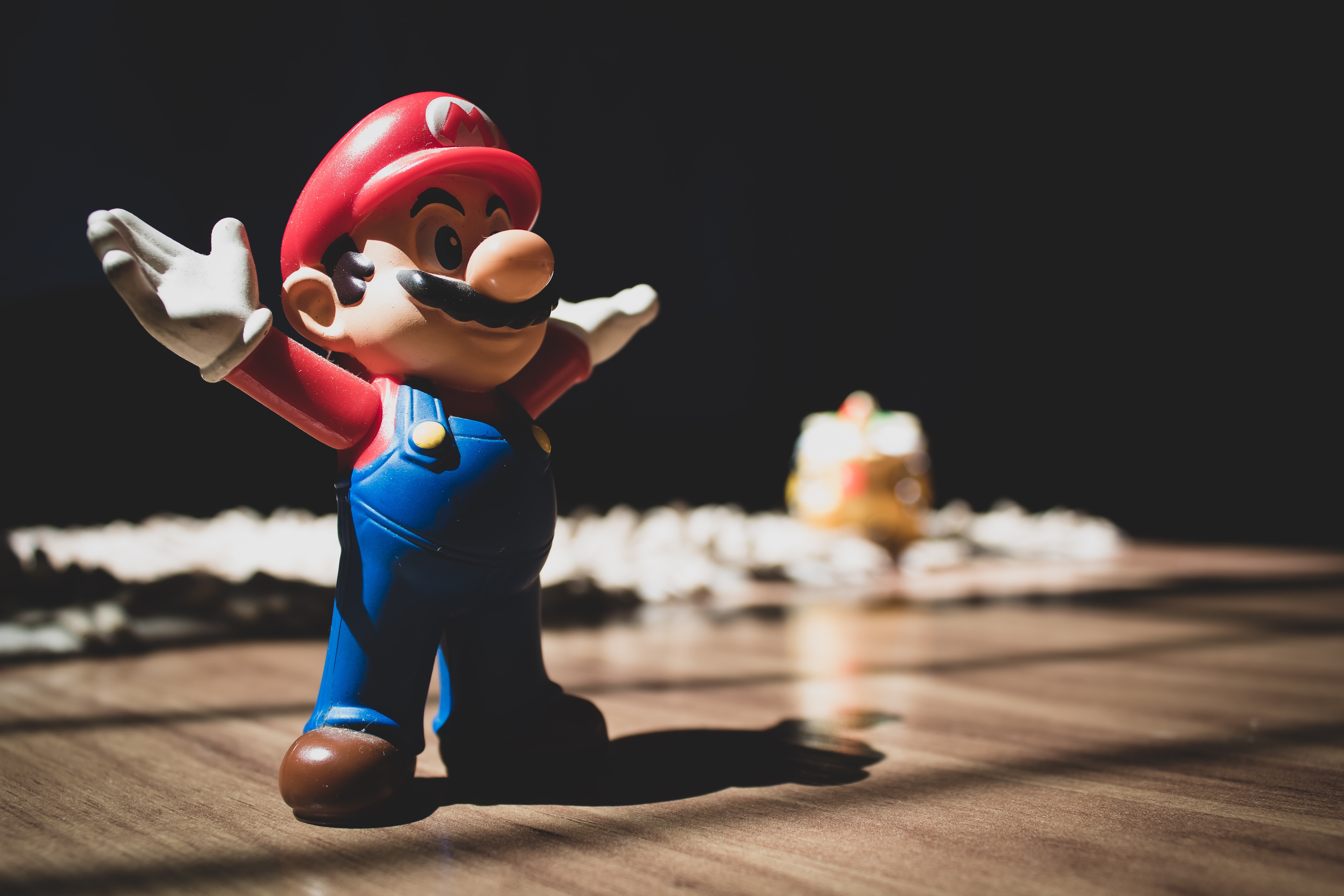 A plastic Super Mario toy set on a table. Mario is standing with his arms out wide and wearing his signature red hat with an M on it, white gloves, red shirt, blue dungarees and brown shoes