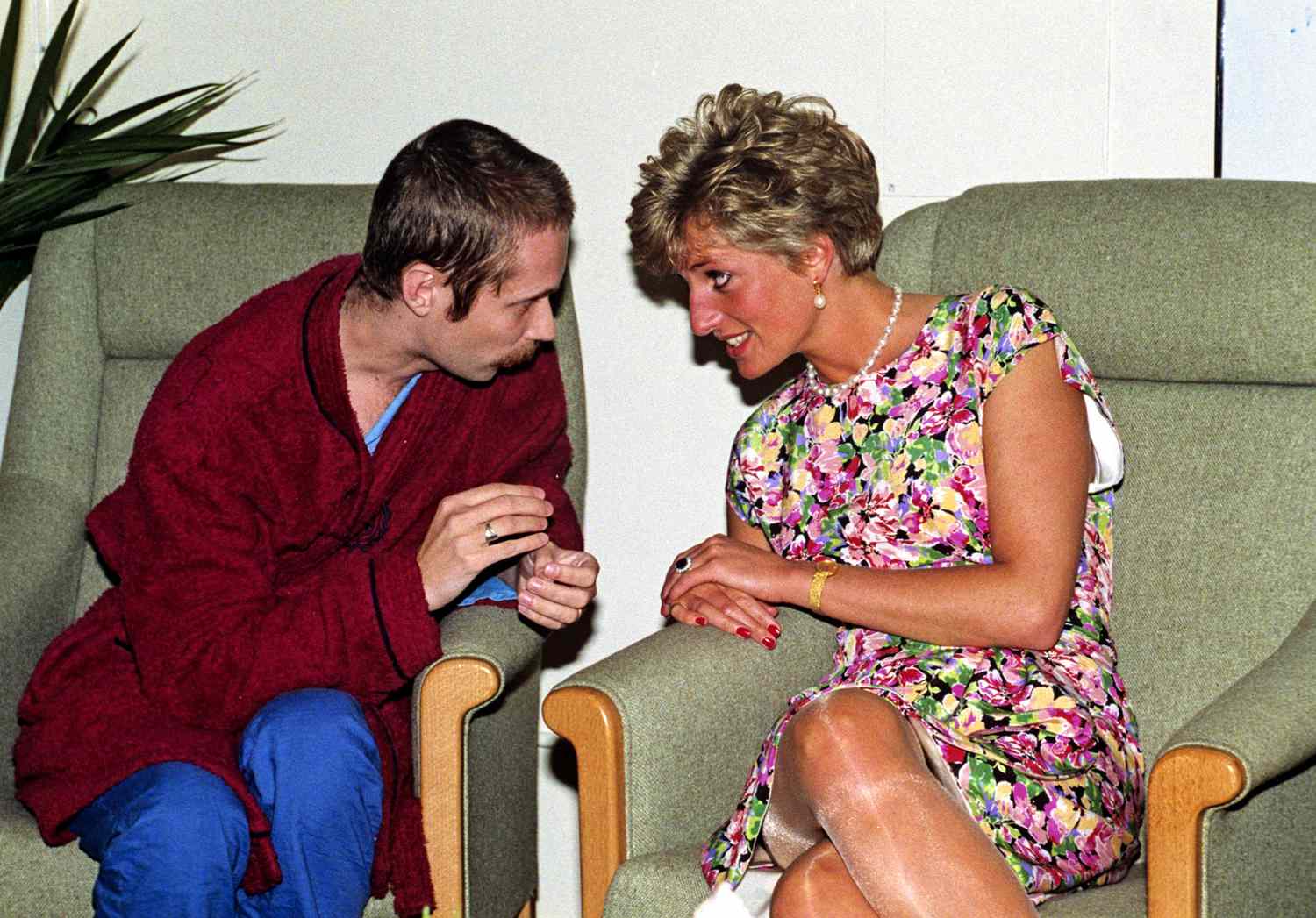 Princess Diana, wearing a floral dress and pearl jewellery, sits in a green armchair next to a man who is wearing a red dressing gown. They are both leaning towards each other, looking into each other eyes engaged in conversation