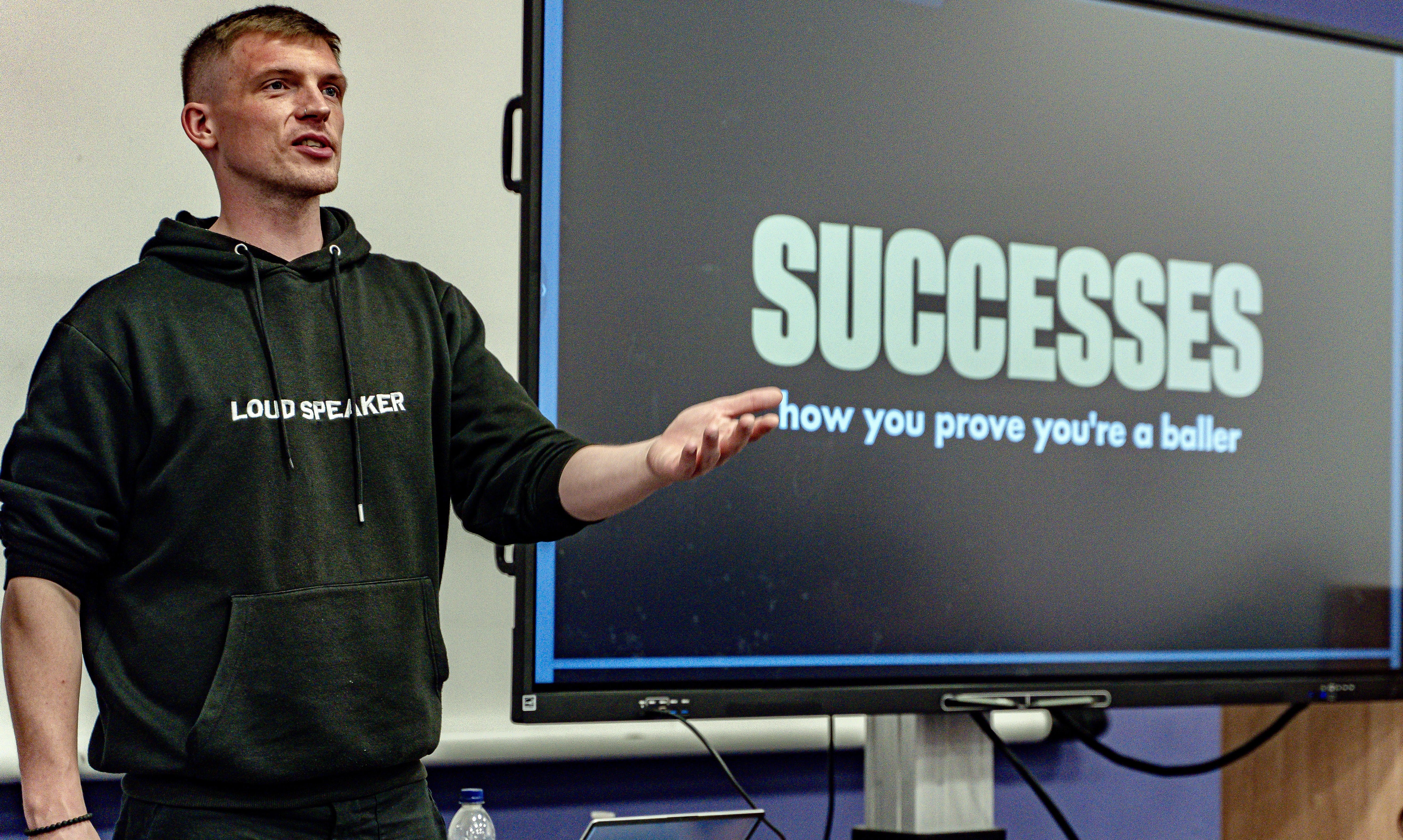 A white man wearing a black hoodie which says Loud Speaker across the middle of it. He is stood next to a screen which says 'Successes, how to prove you're a baller'