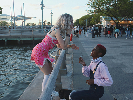 A photo capturing a candid moment along a busy waterfront with a black woman on short red hair, wearing a pink sweater and a black denim jeans, kneeling on one knee  holding up an object facing a white woman with blond hair. The white woman is wearing a short pink side slit dress with white knee-high socks and white sneakers standing and leaning forward with a surprised expression. This gesture suggests a marriage proposal.