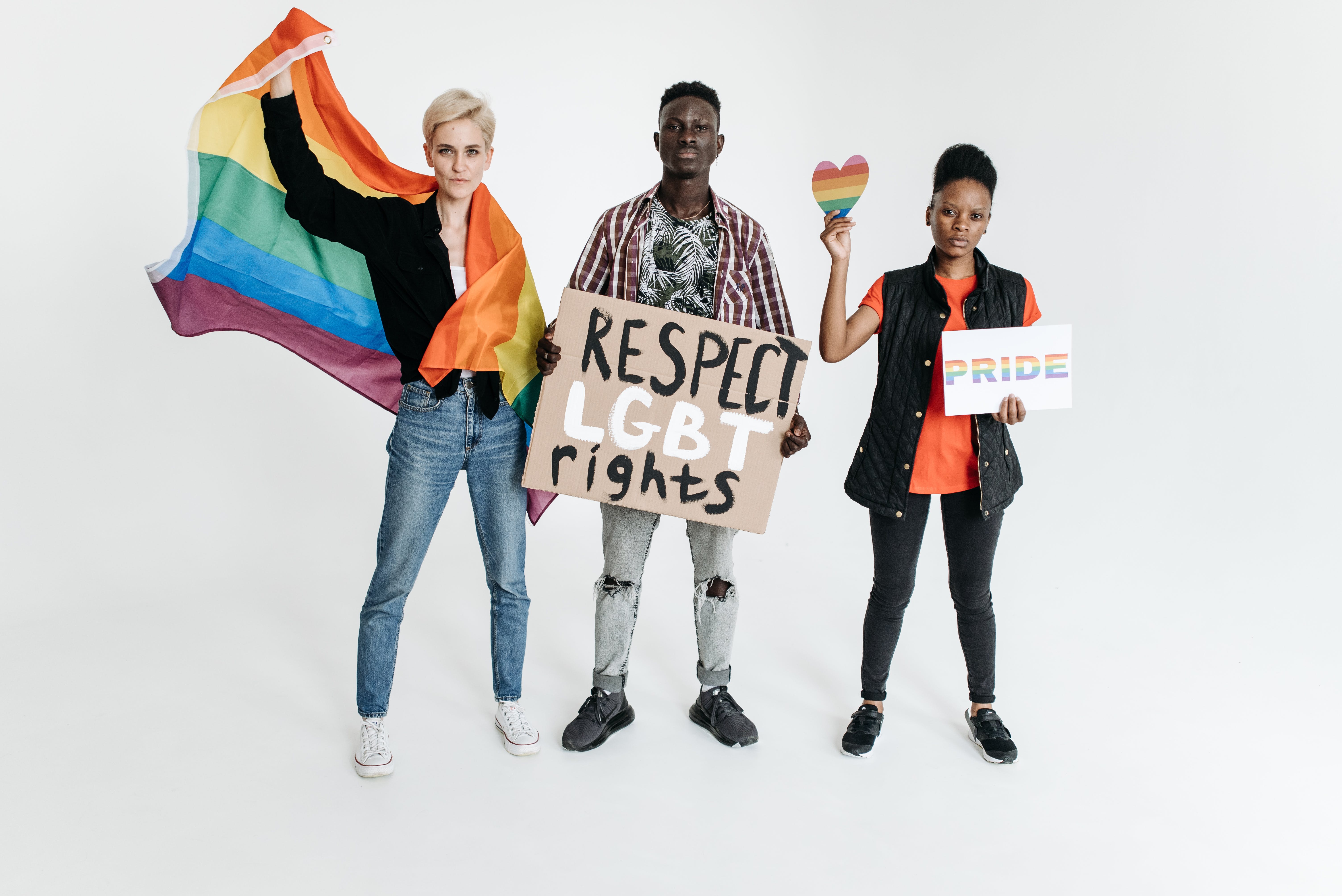 A photo featuring three people standing against a white background advocating for LGBTQ rights. On the left, a white woman with short blonde hair holds a large rainbow flag in the air with her right hand, with it draped over her left shoulder. In the centre, a black man holds a sign reading "Respect LGBT rights," and on the right, a black woman with short black hair holds a small rainbow heart in her right hand and a sign with the word "PRIDE" in rainbow colours in her left hand.