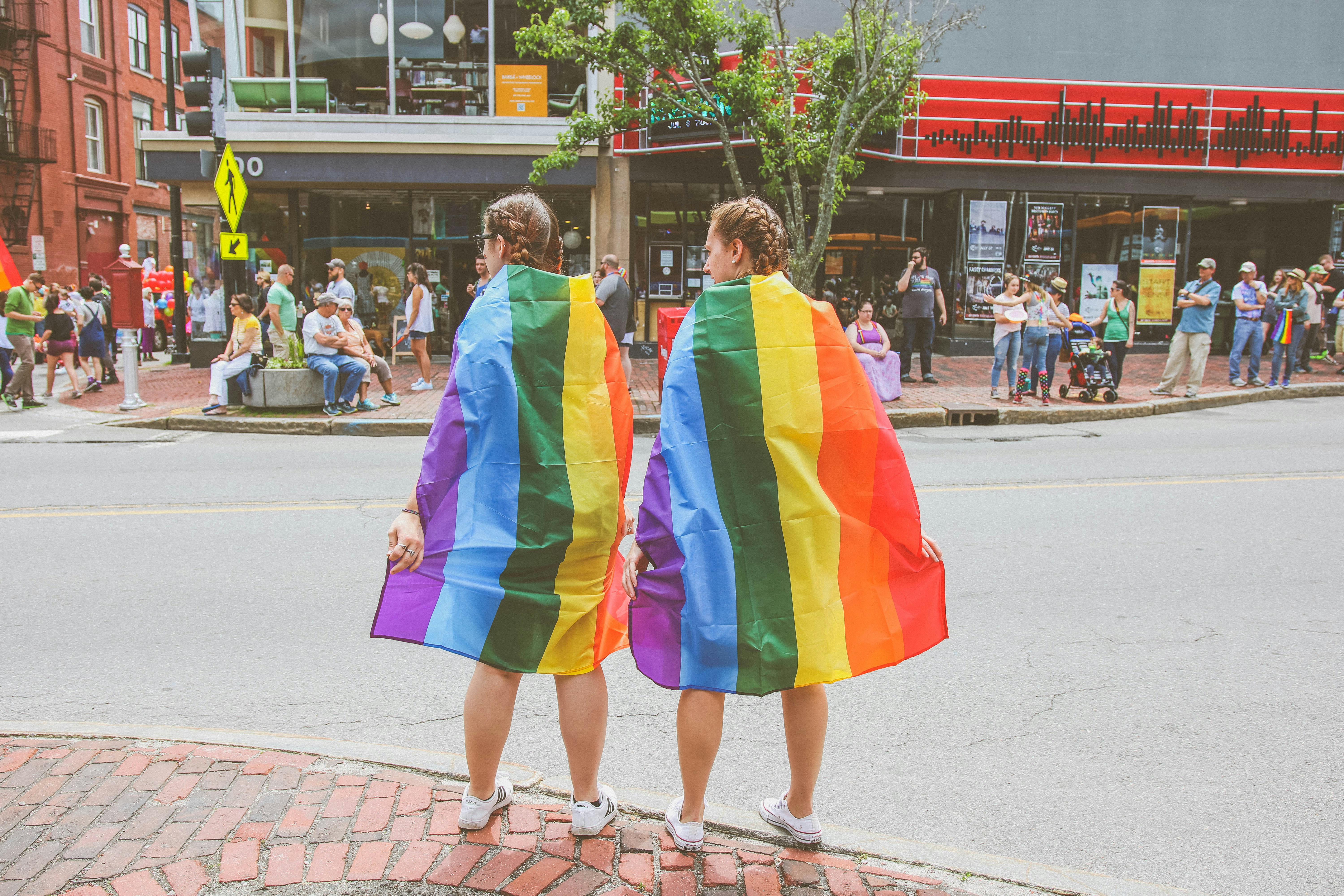 Two women wearing rainbow pride flags as capes stand on a city street, facing away from the camera. The street is bustling with people. The setting appears to be a festive event with a variety of onlookers and participants in the background. 