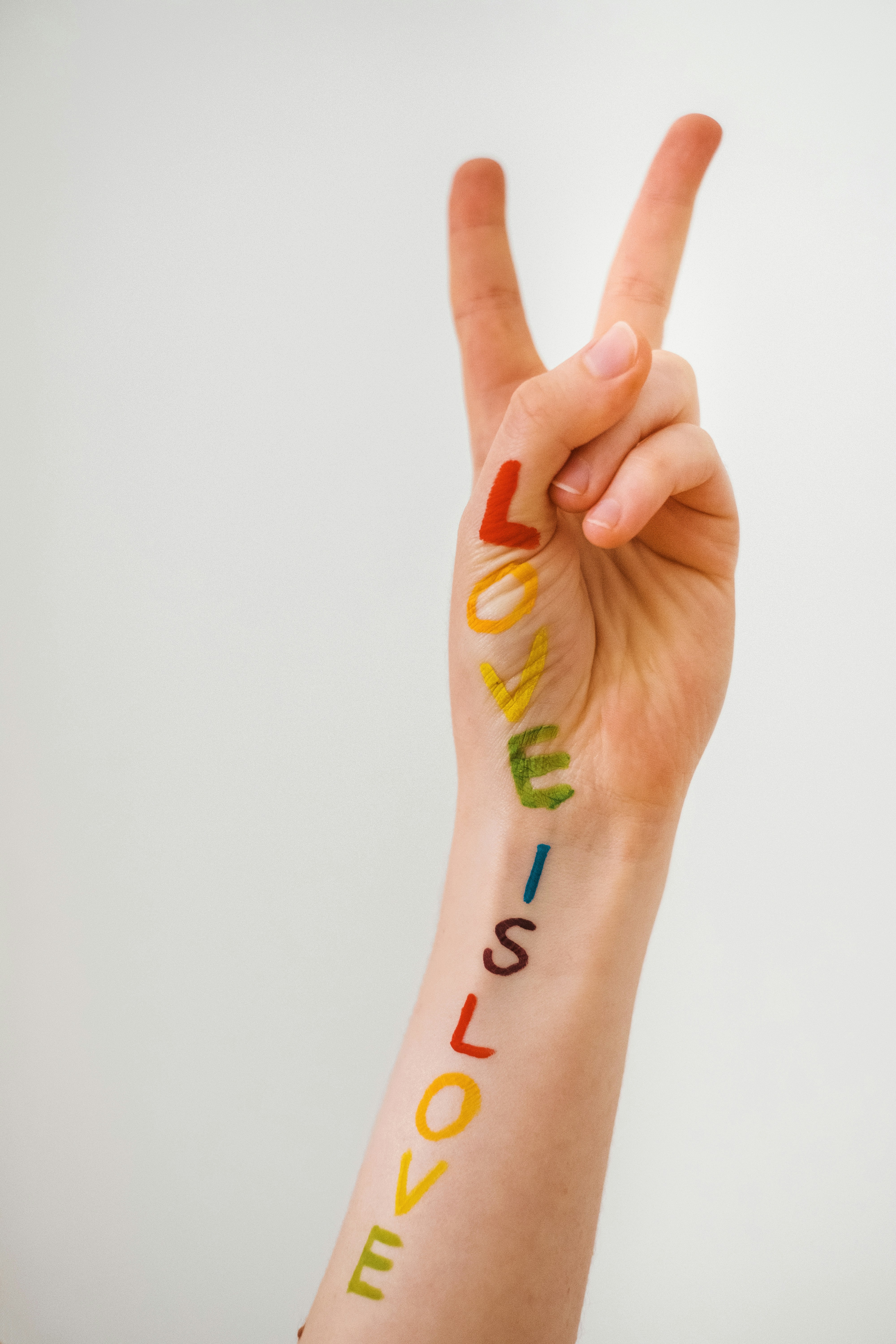 A white arm is raised displaying a peace sign with two fingers in the air, and the phrase "LOVE IS LOVE" is painted in rainbow colours along the forearm. Each letter is in a different colour, corresponding to the stripes of the LGBTQ+ pride flag starting with red at the top and ending with purple at the wrist.