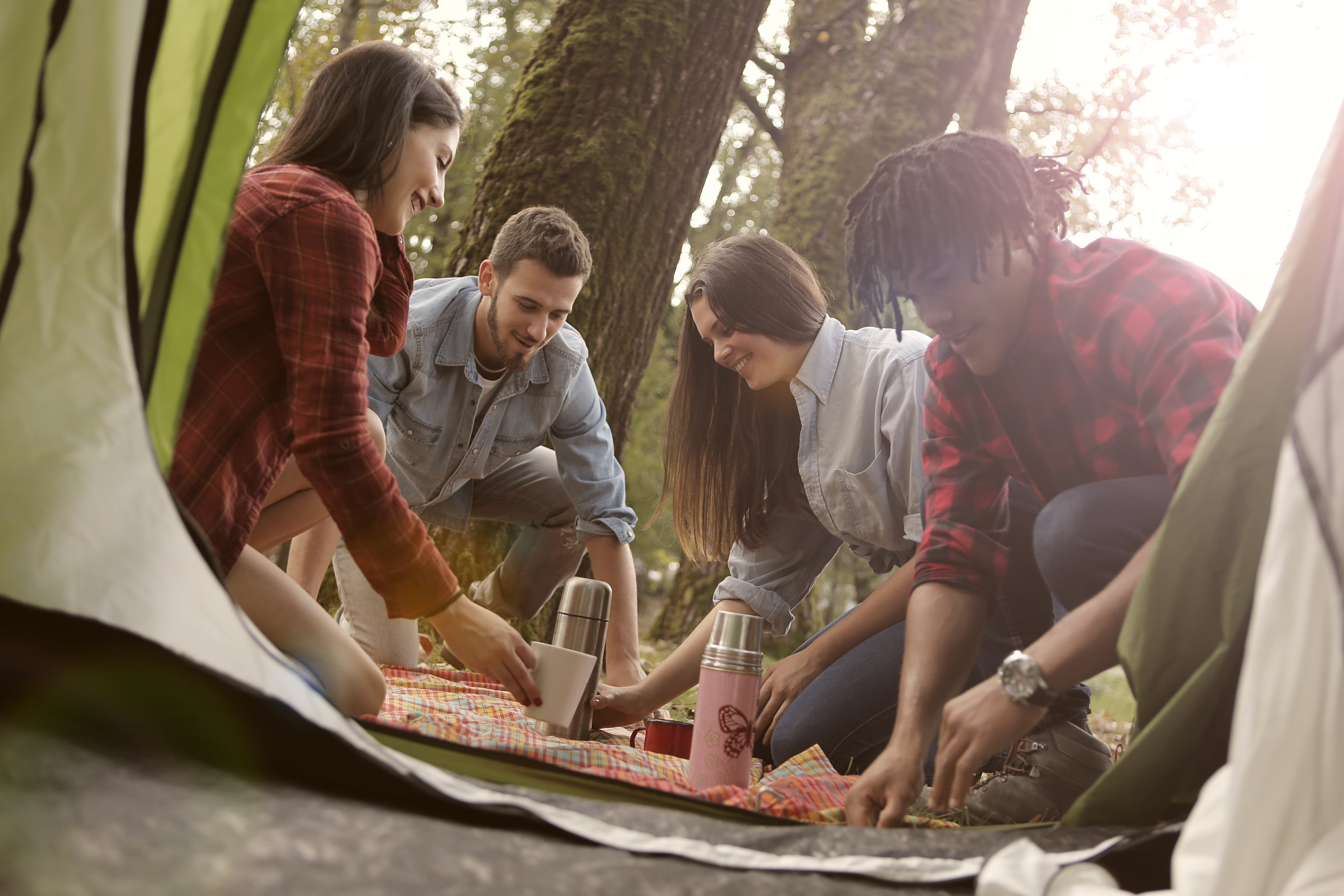 A photo of four young adults enjoying a picnic near a tent in a wooded area. Two women and two men are seen engaging with each other over a colorful picnic blanket spread on the ground. One of the women is reaching for a thermos, while the others appear to be in the midst of conversation and preparing food. The background is softly lit with sunlight filtering through the trees.