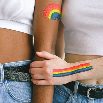 A close-up photo of two faceless women, one black and one white, standing side by side. They are holding hands and wearing denim pants with black belts and white crop tops. Rainbow flags are painted on their skin. On the left, a rainbow flag is painted on an arm with lighter skin, and on the right, the flag is on an arm with darker skin. 