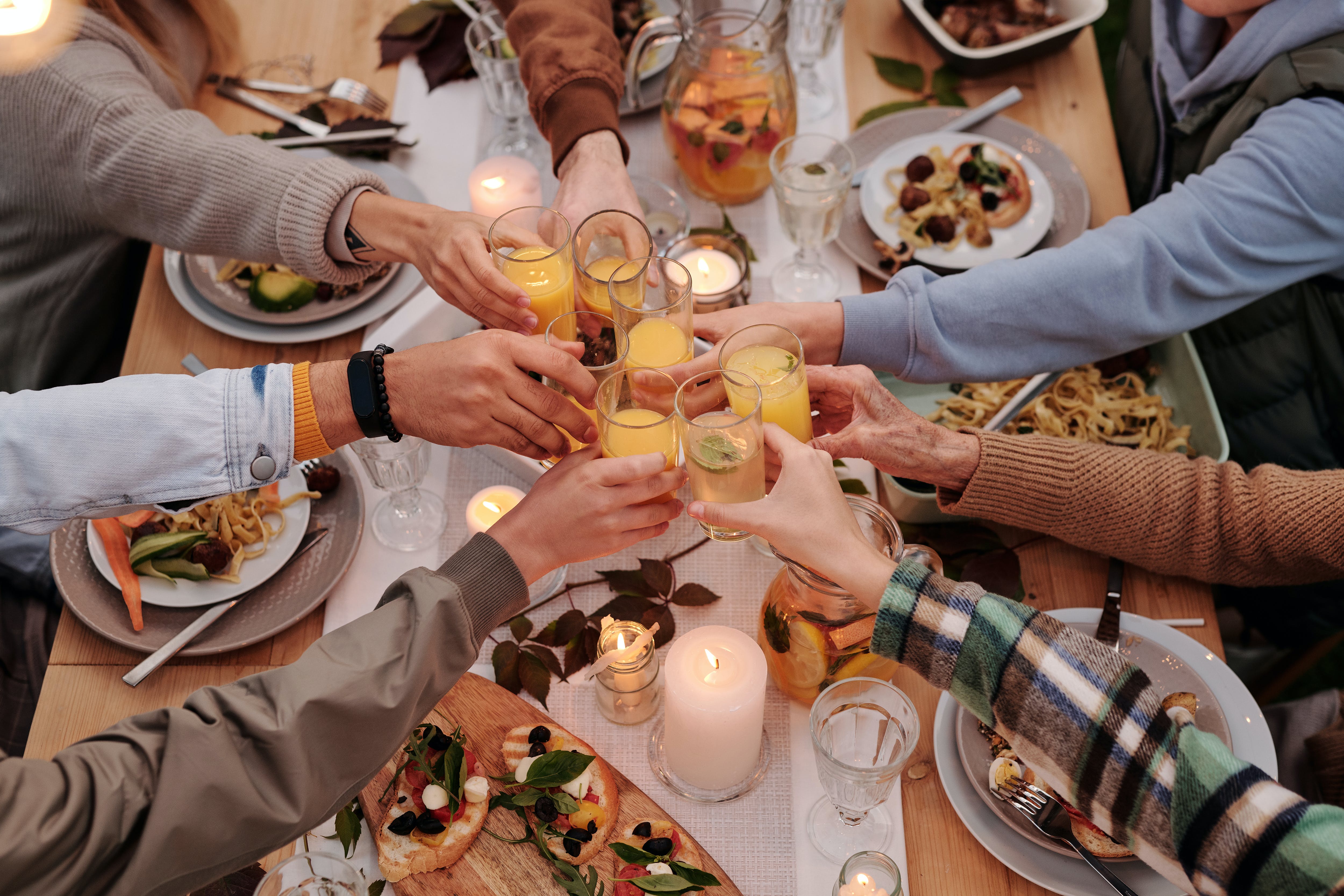 Seven friends sitting around a dinner table with plates of food and candles. Their arms are stretched into the middle of the table raising a toast with their drinks.