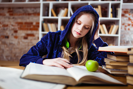 A young woman with her hood up reading an open book at a table. There are stacks of books next to her and an apple.