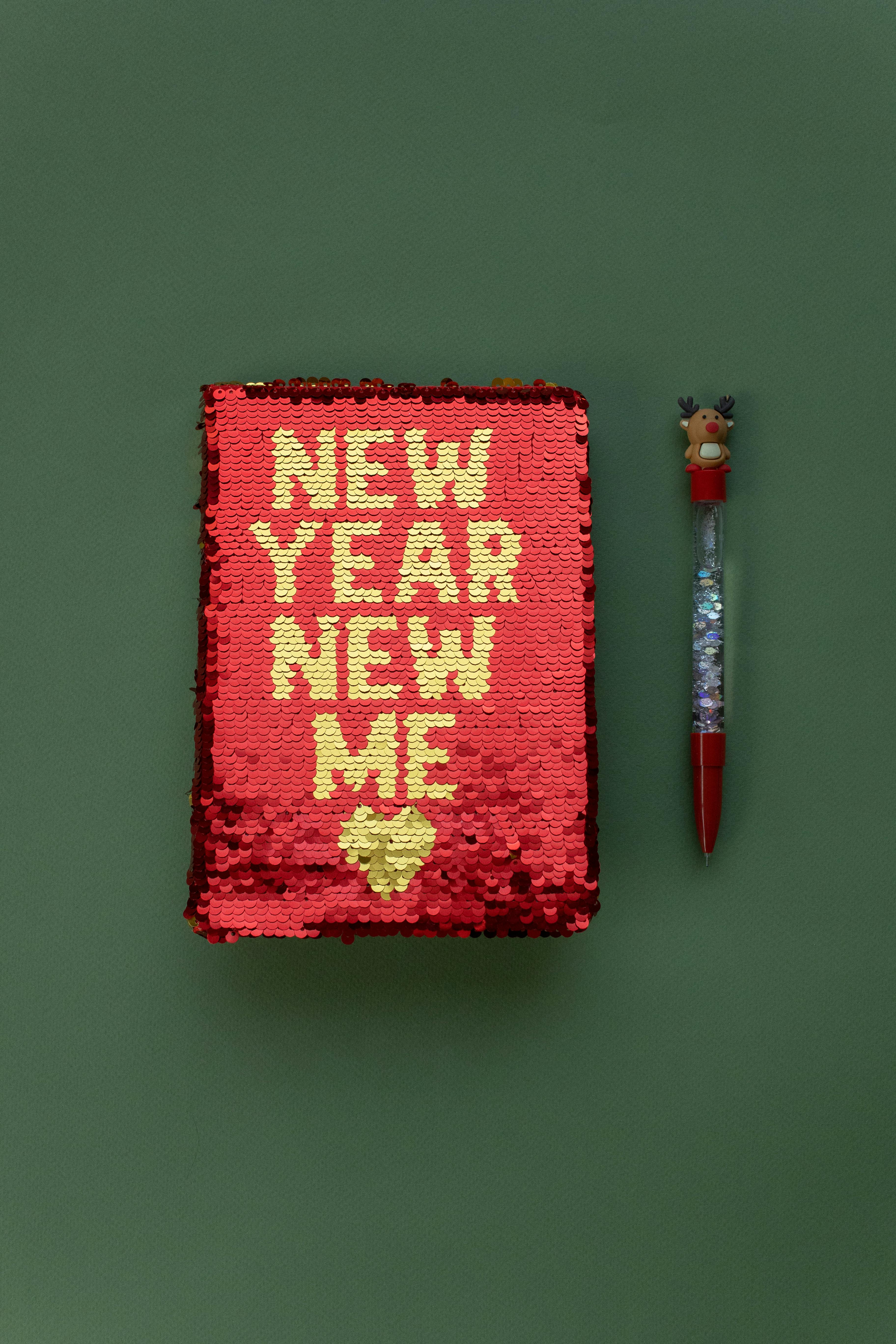 A notebook with a red sequined cover with writing  "NEW YEAR NEW ME" in yellow with a heart shaped drawing underneath the writing. The notebook is on a dark green background. To the right of the notebook, there's a pen decorated with a reindeer figure at the top, including antlers and a red nose, which complements the festive theme. 
