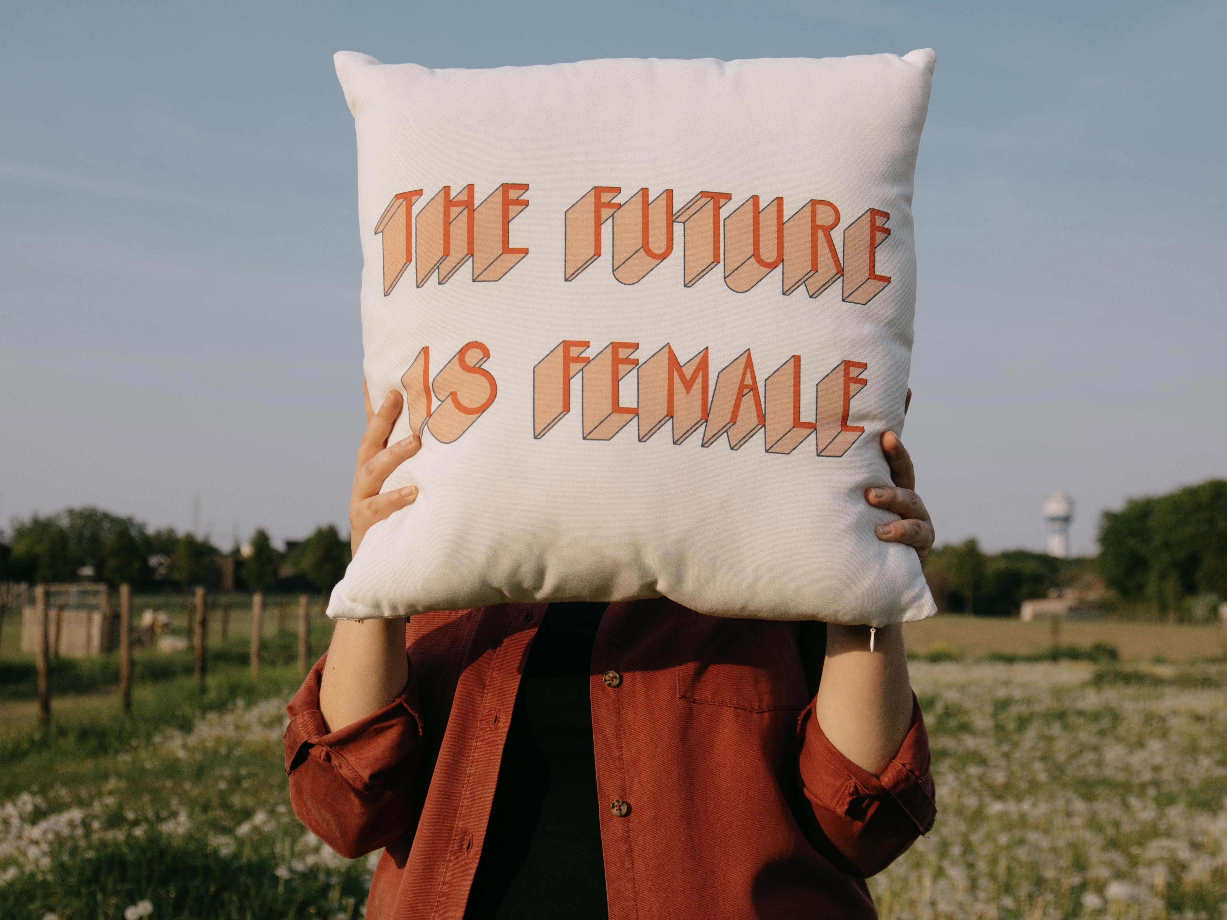 a woman with red shirt and black trousers holding a pillow with inscription saying The future is Female 