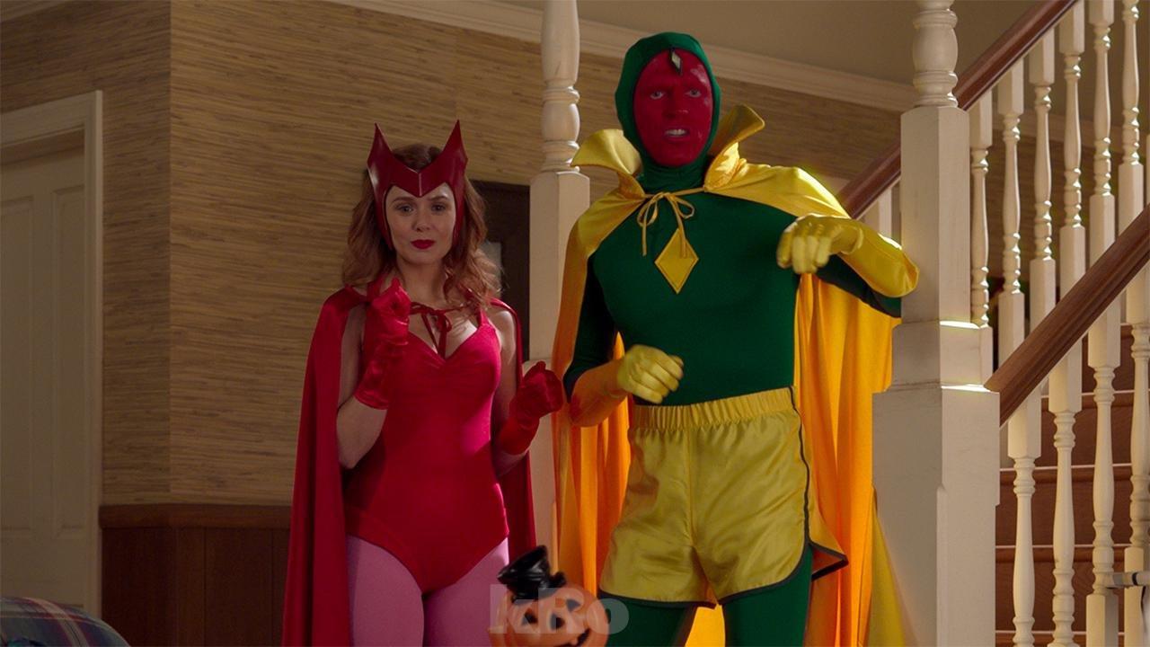 A screenshot from All-New Halloween Spooktacular! the sixth episode of the first season of WandaVision. Wanda is standing to the left wearing a red leotard, cape and gloves, with pink tights and a red crown. Vision is standing to here right wearing a green bodysuit with a yellow cape and trousers, his face is painted red.