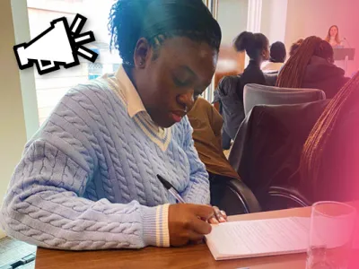 Jemimah, a young black lady is sat at a desk writing notes from a lecture in her notebook.