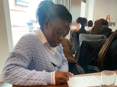 Jemimah, a young black lady is sat at a desk writing notes from a lecture in her notebook.