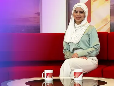 Shamza in a light green blouse and white hijab sitting on a red couch with two mugs on the table.