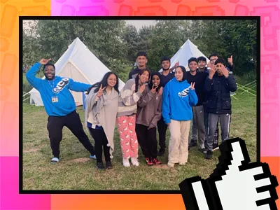 A group of ten young people posing outdoors in front of white tents. They are dressed casually, some wearing hoodies, and appear to be enjoying themselves, with several people making peace signs. The background includes trees and a grassy area, indicating they are likely at a campsite. The image has a colorful border with shades of pink, orange, and yellow, and includes a graphic of a thumbs-up icon in the lower right corner.