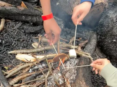 Thee people roasting marshmallows and chocolate digestive biscuits to make smores over a man-made outdoor fire.