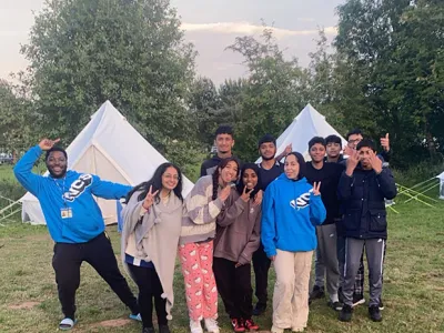 A group of young people posing outdoors in front of white tents. They are dressed casually, some wearing hoodies, and appear to be enjoying themselves, with several people making peace signs. The background includes trees and a grassy area, indicating they are likely at a campsite.