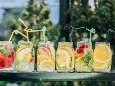 A row of six mason jars filled with fruit-infused water sitting on a table. Each jar contains a variety of fresh fruit slices including watermelon, lime, lemon, orange, and strawberries and mint leaves. There is a straw in each drink.