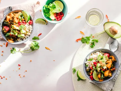 Two bowls filled with vibrant vegetable and rice dishes are placed on a light-colored surface. Each bowl contains a mix of rice, roasted sweet potatoes, avocado slices, diced tomatoes, leafy greens, and lime wedges, garnished with fresh cilantro. 