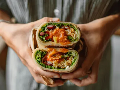 A person holding a vegetable wrap cut in half, showing the colourful and fresh ingredients inside. The sandwich is filled with lettuce, rice, red beans, diced vegetables, and topped with a generous amount of orange sauce