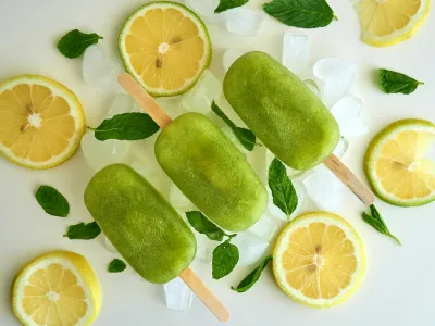 Three green ice lollies are lying on top of a bed of ice cubes, surrounded by slices of yellow lemon and fresh mint leaves.