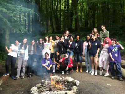 A group of young individuals gathered around a campfire, surrounded by trees in a serene woodland setting.