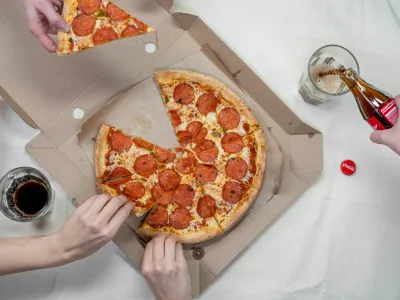 A top-down view of a pepperoni pizza in an open cardboard box, with three hands reaching for slices. One hand pours Coca-Cola from a glass bottle into a glass, while another glass and a Coca-Cola bottle are placed nearby. A red Coca-Cola bottle cap is also visible on the white tablecloth.