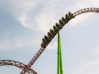 A group of people ride a roller coaster as it ascends a steep track against a clear blue sky. The coaster's red and green structure stands out vividly, highlighting the thrill and height of the ride.