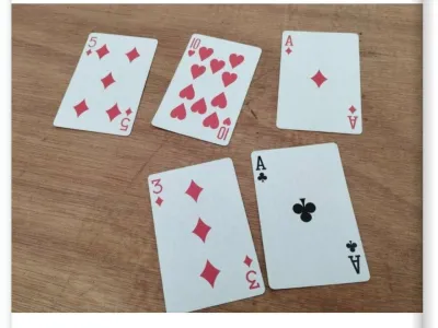 Five playing cards arranged on a wooden surface. The cards shown are the 5 of diamonds, 10 of hearts, Ace of diamonds, 3 of diamonds, and Ace of clubs. The date