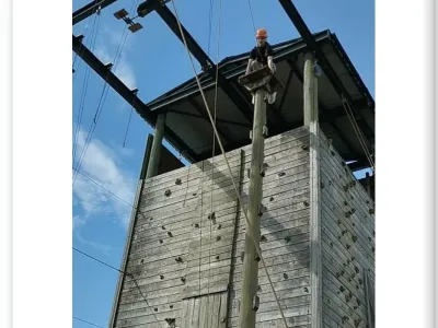 A towering wooden climbing wall against a blue sky. Catriona in an orange helmet stands at top of the structure, which is fitted with safety ropes and climbing holds.ted with safety ropes and climbing holds. Dated "July 30".