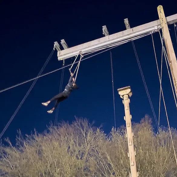 A picture of a person suspended mid-air, holding onto a high ropes course apparatus at night. The person is dressed in dark clothing, and the background shows leafless trees and a dark blue sky. The ropes course consists of cables and wooden poles.