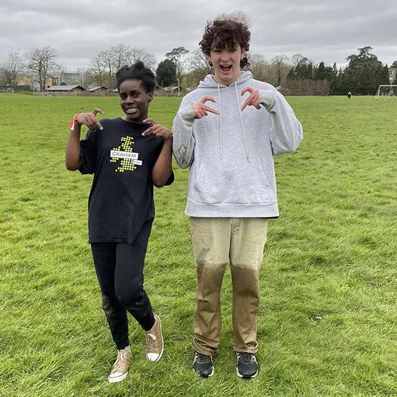 Picture of two young people standing on a grassy field on a cloudy day. The person on the left (a black girl) wears a black T-shirt and black pants, posing with her hands in a playful claw-like gesture. The person on the right (a white boy) wears a grey hoodie and light brown pants with mud stains, also posing with claw-like hands. Trees and a building are visible in the background.