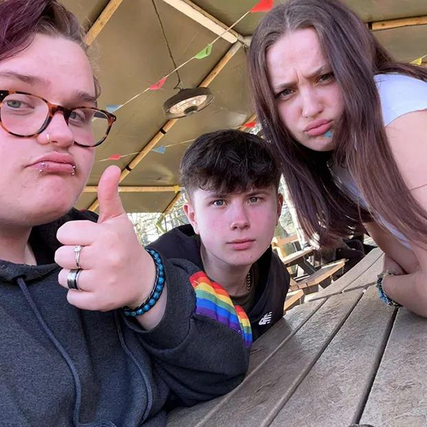 A picture of three young people at a wooden picnic table under a canopy decorated with colourful triangular flags. On the left, a person with glasses, a nose piercing, and short reddish hair gives a thumbs-up while wearing a black hoodie with a rainbow flag on the right sleeve of the hoodie. In the middle, a person with short dark hair wears a . On the right, a person with long brown hair leans on the table and makes a pouty face wearing a white top.